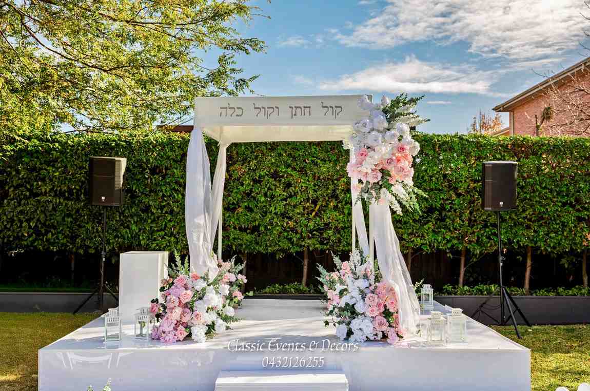 Elegant outdoor Jewish wedding chuppah decorated with white drapes and pink floral arrangements on a garden stage